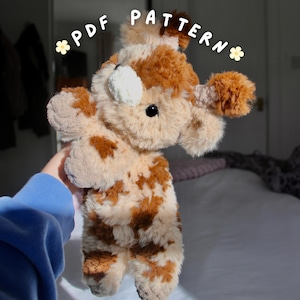 Peut inclure: Une peluche faite à la main en forme de vache. La vache est principalement beige avec des taches et des accents bruns. Le texte "PDF PATTERN" est écrit au-dessus de la vache dans une police fantaisiste. La vache a des yeux noirs et une tache blanche.