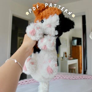 May include: A plush cat toy featuring a calico pattern with patches of orange, black, and white fur. The stuffed animal has pink embroidered paw pads on its feet and dark eyes. The text PDF PATTERN is printed in white above the cat.