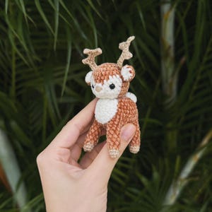 May include: A small, handmade crochet deer figurine. The deer has a brown body, white chest, and tan antlers. It is held in a person's hand against a blurred green background of foliage. The deer is approximately 3 inches tall.