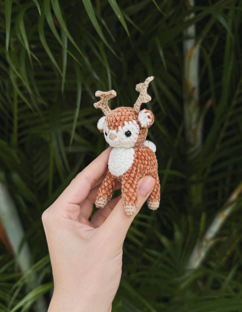 May include: A small, handmade crochet deer figurine. The deer has a brown body, white chest, and tan antlers. It is held in a person's hand against a blurred green background of foliage. The deer is approximately 3 inches tall.