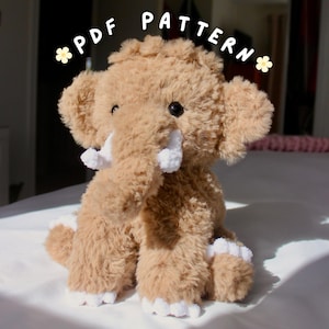 Może przedstawiać: Pluszowy, jasnobrązowy słoń z białymi kłami i stopami. Pluszak ma duże, puszyste uszy i teksturową trąbę. Tekst "PDF PATTERN" jest napisany nad słoniem na biało. Słoń siedzi na białej powierzchni.