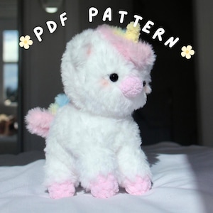 Puede incluir: Un unicornio de peluche blanco con una textura esponjosa, hocico, pezuñas y orejas internas rosas. El unicornio tiene una melena y cola arcoíris, con el texto "PDF PATTERN" encima. El unicornio está sentado sobre una superficie blanca.