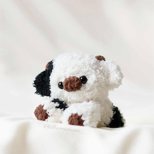 May include: A handmade plush puppy with a white body, black spots, and brown accents. The puppy has black eyes and a brown nose. The plush toy is sitting on a white surface.