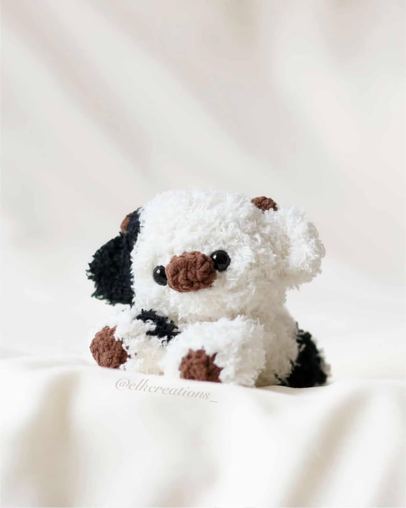 May include: A handmade plush puppy with a white body, black spots, and brown accents. The puppy has black eyes and a brown nose. The plush toy is sitting on a white surface.