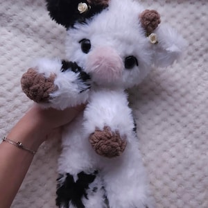 May include: A plush cow toy with a black and white pattern. The toy has brown hooves and ears, and a pink nose. Small white flowers are attached to the ears. The cow is made of soft, fluffy material.