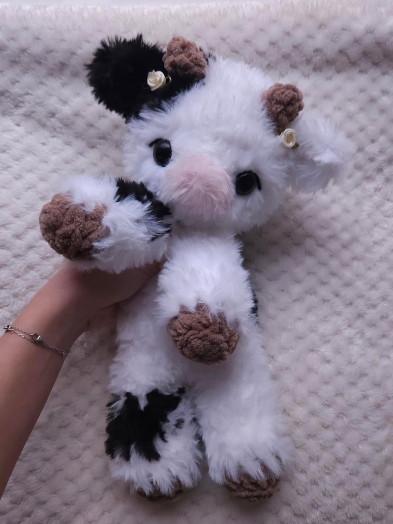 May include: A plush cow toy with a black and white pattern. The toy has brown hooves and ears, and a pink nose. Small white flowers are attached to the ears. The cow is made of soft, fluffy material.