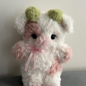 Pu&ograve; includere: Un peluche a forma di mucca, con un corpo bianco e dettagli rosa e verdi. Ha occhi neri, macchie rosa e un fiocco rosa. Le orecchie sono verdi e il design generale &egrave; morbido e soffice.