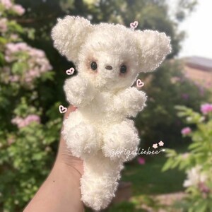 May include: A handmade, fluffy white teddy bear with large, round ears and a small, dark nose and eyes. The bear is made of a soft, textured material and has a cute, cuddly appearance. Small pink hearts are scattered around the bear.