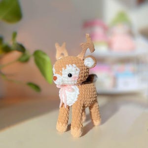 May include: A handmade crochet deer figurine with brown and cream-colored yarn. The deer has a white face, pink cheeks, and a small pink bow. The antlers are a lighter brown. The deer is standing on four legs.