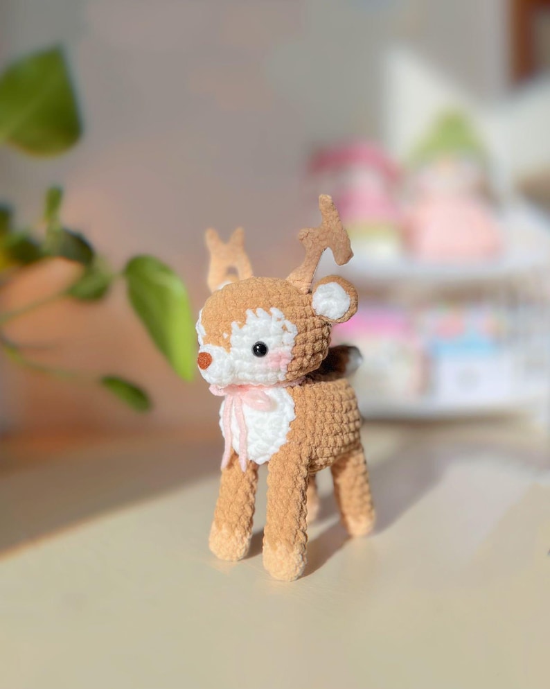 May include: A handmade crochet deer figurine with brown and cream-colored yarn. The deer has a white face, pink cheeks, and a small pink bow. The antlers are a lighter brown. The deer is standing on four legs.