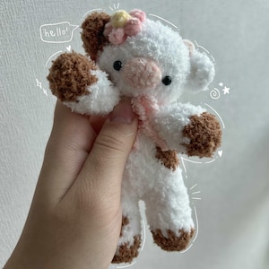 May include: A handmade plush cow toy with white fur and brown spots on its hooves, ears, and body. It has a pink scarf and a flower on its head. The word "hello!" is written in a speech bubble.