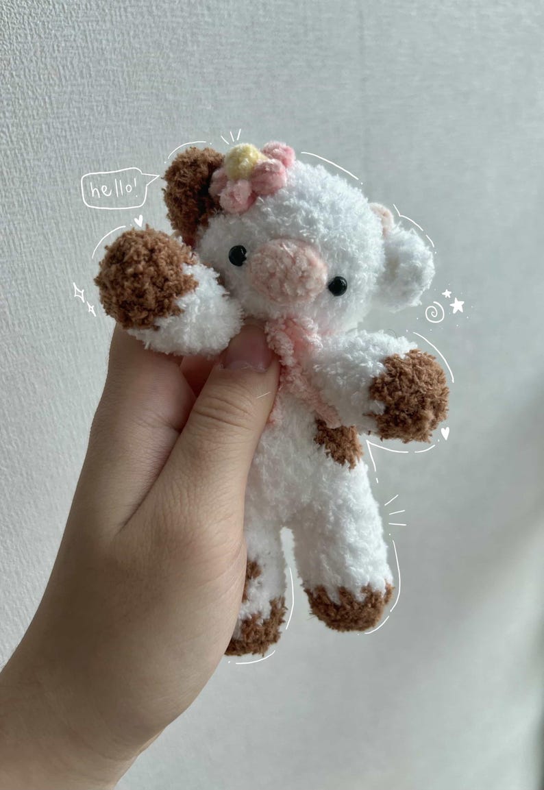 May include: A handmade plush cow toy with white fur and brown spots on its hooves, ears, and body. It has a pink scarf and a flower on its head. The word "hello!" is written in a speech bubble.