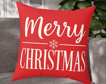 May include: A red square pillow with the words "Merry Christmas" in white script and block letters. A white snowflake graphic is centered between the words. The pillow is resting on a gray sofa.