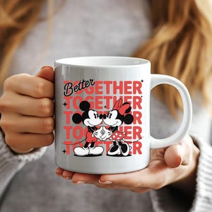 Op de afbeelding: Witte keramische mok met Mickey en Minnie Mouse in een romantische pose. De mok heeft de woorden "Better Together" in rood en zwart. De mok wordt door een persoon vastgehouden.