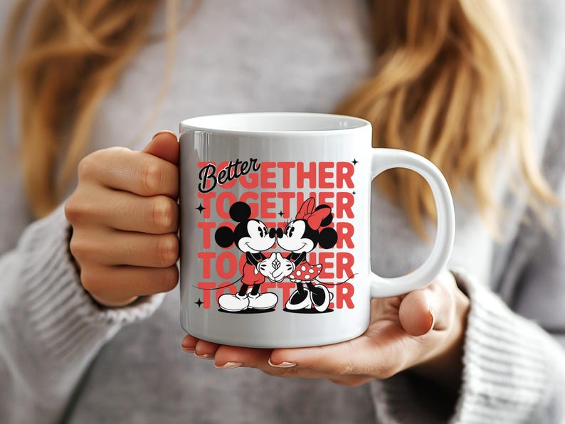 Op de afbeelding: Witte keramische mok met Mickey en Minnie Mouse in een romantische pose. De mok heeft de woorden "Better Together" in rood en zwart. De mok wordt door een persoon vastgehouden.