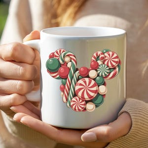Puede incluir: Taza de cer&aacute;mica blanca con un dise&ntilde;o de caramelos de colores en forma de cabeza de rat&oacute;n. El dise&ntilde;o incluye bastones de caramelo a rayas rojas y blancas, caramelos redondos verdes y rojos y caramelos en espiral rojos y blancos. La taza es sostenida por una persona.