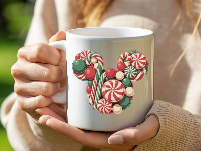Puede incluir: Taza de cer&aacute;mica blanca con un dise&ntilde;o de caramelos de colores en forma de cabeza de rat&oacute;n. El dise&ntilde;o incluye bastones de caramelo a rayas rojas y blancas, caramelos redondos verdes y rojos y caramelos en espiral rojos y blancos. La taza es sostenida por una persona.