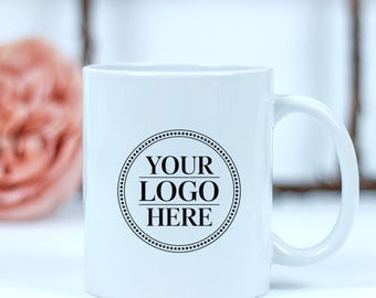 May include: A white ceramic mug with a curved handle. The mug features a black circular design with the text "YOUR LOGO HERE" in a bold, sans-serif font. The mug is designed for custom branding.