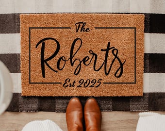 May include: A rectangular doormat with the text "The Roberts Est 2025" in black script. The mat is brown and framed with a black border. It rests on a black and white checkered rug. Brown boots and blue jeans are visible.