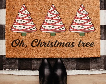 May include: A rectangular doormat with three illustrated Christmas trees in the center. The trees are white with red ribbon and green accents. The text "Oh, Christmas tree" is below the trees. The mat is on a black and white checkered rug.