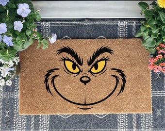 May include: A rectangular doormat featuring the Grinch's face with yellow eyes and a mischievous grin. The mat is made of a natural brown material and is placed on a patterned rug, with potted flowers on either side.