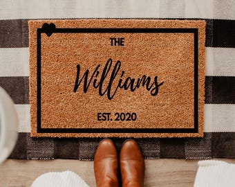 May include: A rectangular doormat with the text "THE Williams EST. 2020" in black script, framed by a black border. The mat is brown and sits on a black and white checkered rug. Brown boots are visible.