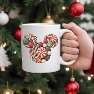 Puede incluir: Taza de cer&aacute;mica blanca con un dise&ntilde;o de caramelos en forma de cabeza de rat&oacute;n. El dise&ntilde;o incluye caramelos rojos, verdes y blancos. La taza se sostiene frente a un &aacute;rbol de Navidad.
