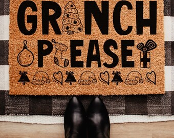 May include: A rectangular doormat with the words "GRINCH PLEASE" in black, along with Christmas-themed illustrations. The mat is brown with a textured surface, and the background includes a black and white checkered pattern and a wooden floor.
