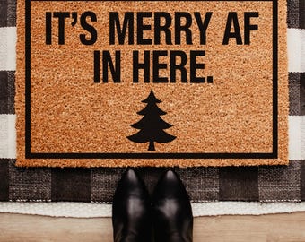May include: A rectangular doormat with the text "IT'S MERRY AF IN HERE" in black, along with a black Christmas tree graphic. The mat is brown with a black border, and is placed on a black and white checkered rug. Black boots and blue jeans are visible.