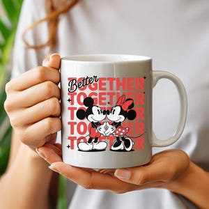 Op de afbeelding: Witte keramische mok met Mickey en Minnie Mouse die elkaar omarmen. De mok heeft de tekst "Better Together" in rode letters. De mok wordt door een persoon vastgehouden.