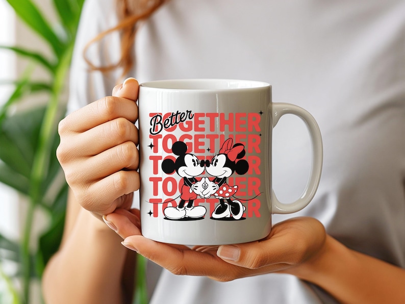 Op de afbeelding: Witte keramische mok met Mickey en Minnie Mouse die elkaar omarmen. De mok heeft de tekst "Better Together" in rode letters. De mok wordt door een persoon vastgehouden.