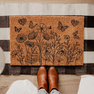 May include: A rectangular doormat with a floral and butterfly design. The mat is made of a natural brown material and features black line art of flowers, leaves, and butterflies. The mat is placed on a black and white checkered rug.