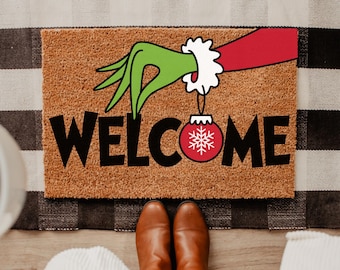 May include: A brown coir doormat with the word "WELCOME" in black. The design features a green hand holding a red Christmas ornament with a white snowflake. The doormat is placed on a black and white checkered rug.