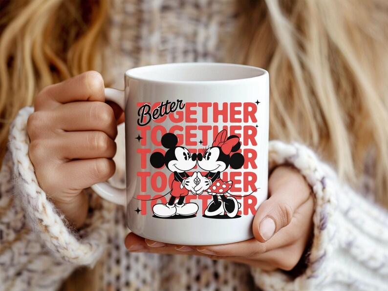 Op de afbeelding: Witte keramische mok met Mickey en Minnie Mouse die elkaar omarmen, met de woorden "Better Together" in het rood. De mok heeft een gebogen handvat en wordt vastgehouden door een persoon die een witte trui draagt.