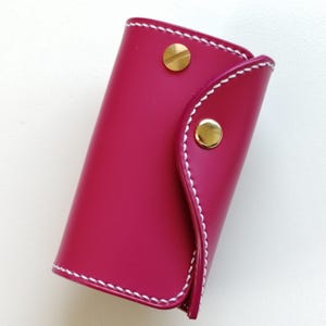 Può includere: Un astuccio portachiavi color magenta vivace con finitura liscia e cuciture bianche a contrasto lungo i bordi. L'accessorio è fissato con due bottoni a pressione in metallo dorato lucido, uno sulla patta superiore e uno sul lato.