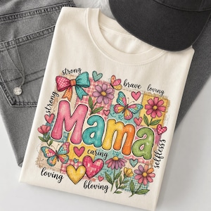Pode incluir: Camiseta creme com a palavra "Mama" em letras coloridas e costuradas. O design inclui borboletas, corações, flores e as palavras "forte", "corajosa", "amorosa", "atenciosa" e "altruísta". Um boné de beisebol preto e jeans cinza estão no fundo.