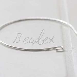 Könnte beinhalten: Ein silbernes Armreif-Armband mit einem einfachen, offenen Design. Das Armband besteht aus einem einzigen, dünnen Stück Metall, das zu einem Kreis gebogen ist. Die Enden des Armbands sind mit einem kleinen, gebogenen Haken verbunden.