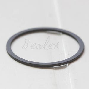 May include: A dark grey, circular metal ring with the word "Beadex" in cursive script. The ring is set against a blurred, light background, highlighting its simple, minimalist design. The ring appears to be a jewellery component or finding.