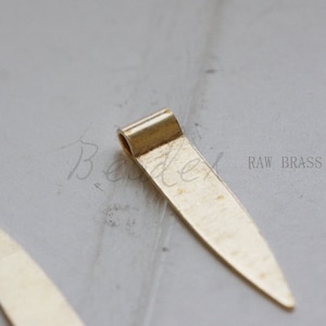 Puede incluir: Un colgante de latón en bruto con una punta puntiaguda y un bucle en la parte superior. RAW BRASS está escrito en el fondo.