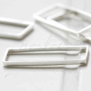 May include: Close-up of several silver rectangular jewelry findings. The findings are open-frame rectangles with a smooth, polished finish. The pieces are arranged on a light-colored surface, with soft lighting highlighting their metallic sheen. These findings are ideal for jewelry making.