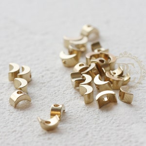 Pode incluir: Uma coleção de pequenos achados de joalheria em metal dourado, cada um em forma de meia-lua. Os achados têm uma superfície lisa e polida e um pequeno orifício para fixação. As peças estão espalhadas sobre uma superfície branca.