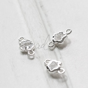 Puede incluir: Tres abalorios conectores de plata en forma de flor con detalles de cristal transparente. Cada abalorio tiene dos lazos para sujetar a las joyas.