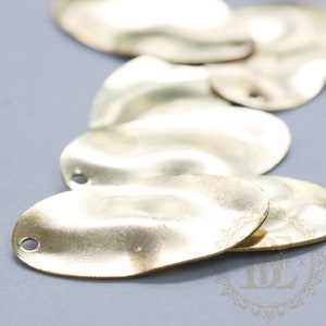 Op de afbeelding: Een close-up van verschillende golvende, ovale metalen bedels met een gat aan de bovenkant. De bedels zijn licht goud van kleur en hebben een licht getextureerd oppervlak.