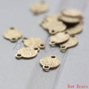 May include: A group of small, round, brass charms with two jump rings. The charms have a textured surface and are scattered on a light gray surface. Raw Brass.