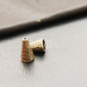 Puede incluir: Dos cuentas de metal de color dorado en forma de cono con textura martillada.