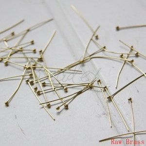 May include: A pile of gold-colored head pins with round heads. The pins are scattered on a white surface. Raw Brass.