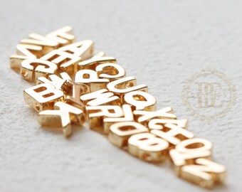 May include: A pile of gold-colored metal letter beads. The letters are in a random order and are all uppercase.
