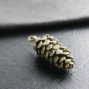 Può includere: Un ciondolo a forma di pigna di colore bronzo con un anello per appenderlo. Il ciondolo è dettagliato con squame individuali e ha un aspetto rustico e naturale.