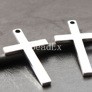 Op de afbeelding: Twee zilverkleurige metalen kruisbedels met een gat aan de bovenkant om op te hangen. De kruisen zijn eenvoudig en elegant, met een licht getextureerd oppervlak.