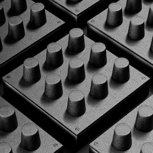 Puede incluir: Imagen monocromática de varios paneles cuadrados grises, cada uno con una cuadrícula de perillas cilíndricas. Las perillas están espaciadas uniformemente y tienen una superficie ligeramente texturizada. Los paneles están dispuestos en un patrón repetitivo.